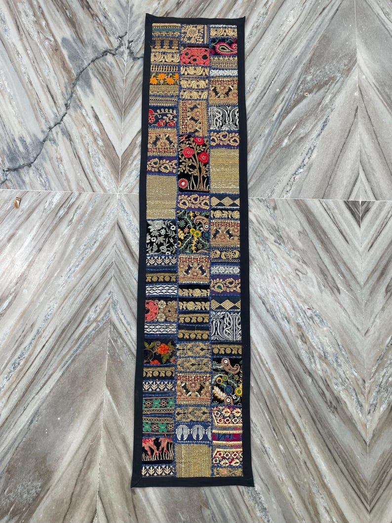 May include: A long, rectangular patchwork textile with a black border. The patchwork design features a variety of colorful, embroidered squares with floral and geometric patterns. The textile is likely a table runner or wall hanging.