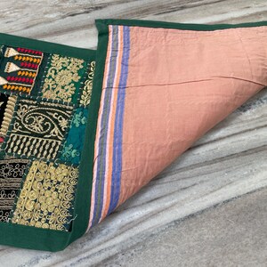 May include: A decorative patchwork textile with a green border. The top features a variety of embroidered patches in shades of green, gold, red, and black. The underside is a solid peach color with striped accents.