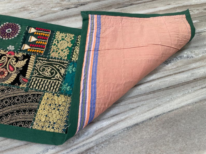 May include: A decorative patchwork textile with a green border. The top features a variety of embroidered patches in shades of green, gold, red, and black. The underside is a solid peach color with striped accents.