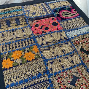 May include: A patchwork textile with a black border. The design features various embroidered squares in shades of blue, gold, red, and orange. Intricate patterns and floral motifs are visible throughout the piece, creating a vibrant and detailed aesthetic.