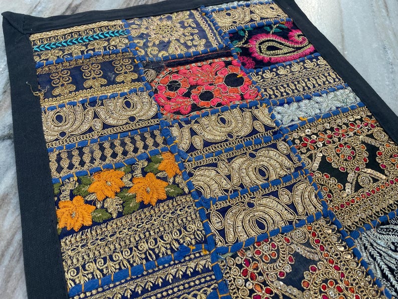 May include: A patchwork textile with a black border. The design features various embroidered squares in shades of blue, gold, red, and orange. Intricate patterns and floral motifs are visible throughout the piece, creating a vibrant and detailed aesthetic.