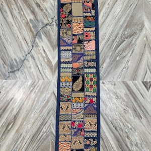 May include: A rectangular, patchwork table runner with a navy blue border. The patchwork design features various patterns and colors, including floral, paisley, and geometric motifs. The runner is likely made of fabric and is designed to decorate a table.