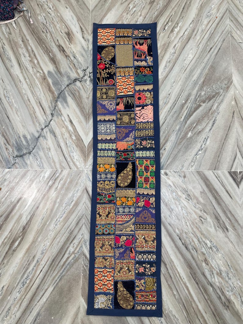 May include: A rectangular, patchwork table runner with a navy blue border. The patchwork design features various patterns and colors, including floral, paisley, and geometric motifs. The runner is likely made of fabric and is designed to decorate a table.