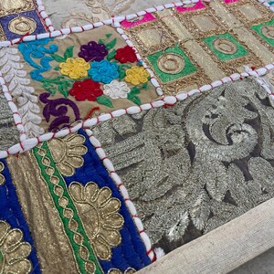 May include: Close-up of a patchwork textile with embroidered squares. The squares feature floral and geometric designs in gold, blue, green, red, and beige. The edges are stitched with white thread and red accents.