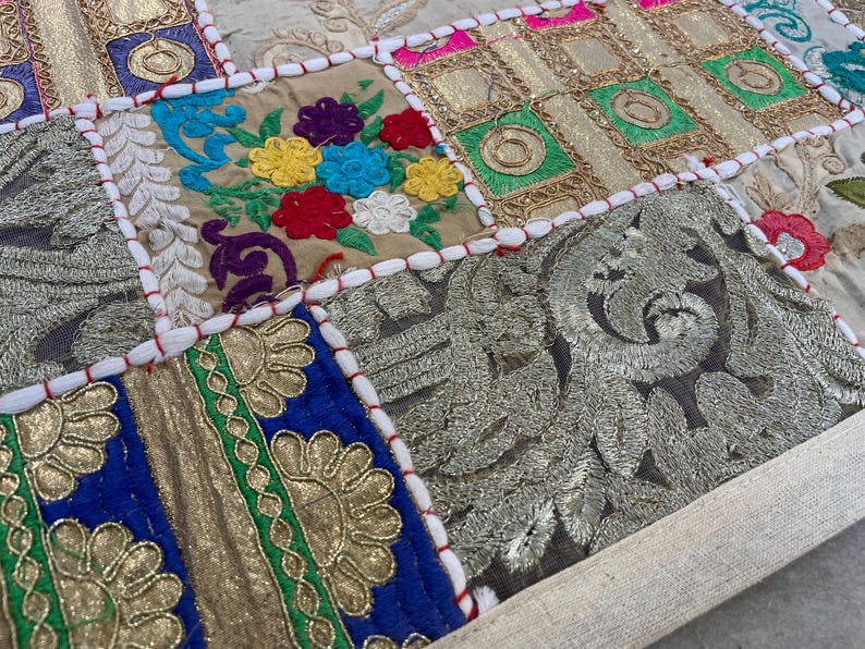 May include: Close-up of a patchwork textile with embroidered squares. The squares feature floral and geometric designs in gold, blue, green, red, and beige. The edges are stitched with white thread and red accents.