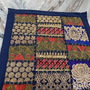 May include: A patchwork textile with a navy blue border. The design features embroidered squares in gold, red, and blue. The squares have floral and geometric patterns. The textile is likely a decorative item, such as a pillow cover or wall hanging.