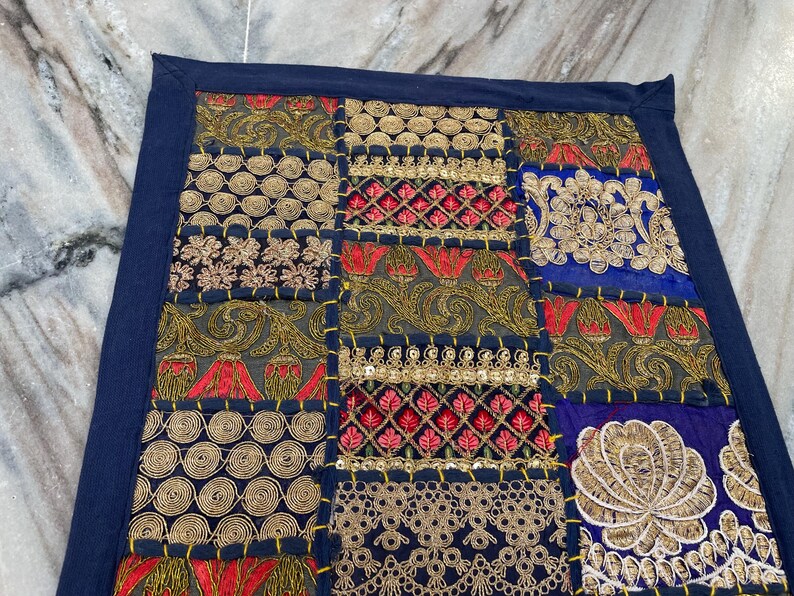 May include: A patchwork textile with a navy blue border. The design features embroidered squares in gold, red, and blue. The squares have floral and geometric patterns. The textile is likely a decorative item, such as a pillow cover or wall hanging.