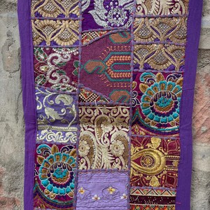 May include: A decorative textile with a patchwork design. The piece features various rectangular panels in shades of purple, gold, and teal, with intricate embroidered patterns. The edges are trimmed with a solid purple border.
