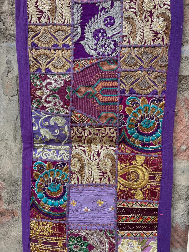 May include: A decorative textile with a patchwork design. The piece features various rectangular panels in shades of purple, gold, and teal, with intricate embroidered patterns. The edges are trimmed with a solid purple border.