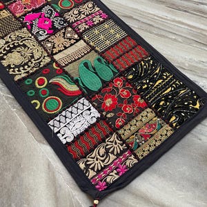 以下が含まれることがあります： 黒い縁取りの長方形パッチワークテーブルランナー。赤、ピンク、金、緑などの鮮やかな色の刺繍が施された正方形が特徴です。デザインには花柄や幾何学模様が含まれています。角にはタッセルが付いています。長さ約150cm。