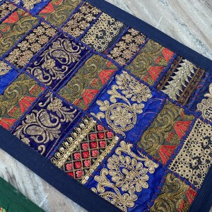 May include: A rectangular, patchwork textile with a dark blue border. The design features squares of blue, gold, red, and black fabrics with embroidered patterns. The textile is likely a decorative item, such as a table runner or wall hanging.