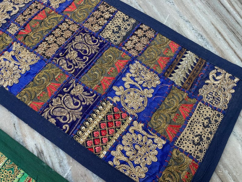 May include: A rectangular, patchwork textile with a dark blue border. The design features squares of blue, gold, red, and black fabrics with embroidered patterns. The textile is likely a decorative item, such as a table runner or wall hanging.