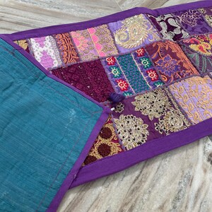 May include: A patchwork table runner with a teal backing and a purple border. The patchwork design features various fabrics in shades of purple, pink, and gold, with intricate embroidered details. The runner is approximately 12 inches wide.