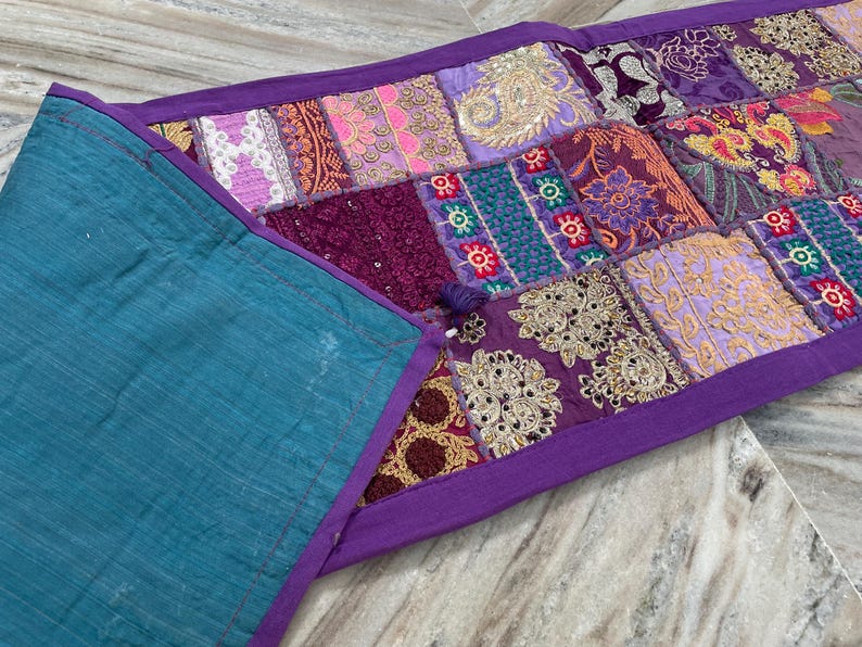 May include: A patchwork table runner with a teal backing and a purple border. The patchwork design features various fabrics in shades of purple, pink, and gold, with intricate embroidered details. The runner is approximately 12 inches wide.