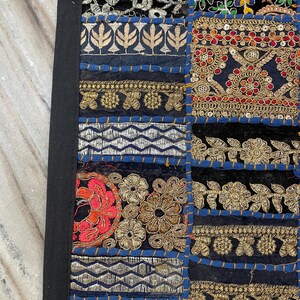 May include: Close-up of a patchwork textile with a black border. The design features various embroidered squares in black, blue, gold, red, and silver. The squares have floral and geometric patterns, with intricate stitching and embellishments.