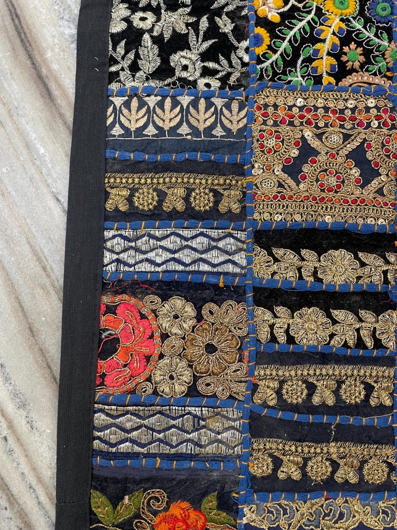 May include: Close-up of a patchwork textile with a black border. The design features various embroidered squares in black, blue, gold, red, and silver. The squares have floral and geometric patterns, with intricate stitching and embellishments.