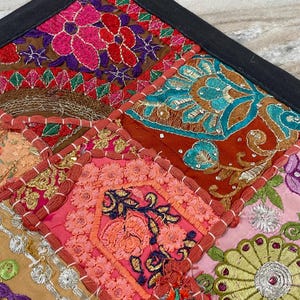 May include: Close-up of a colorful patchwork textile with a black border. The design features embroidered squares in shades of pink, orange, teal, and brown, with floral and geometric patterns. The textile is likely a decorative item, such as a pillow cover or wall hanging.