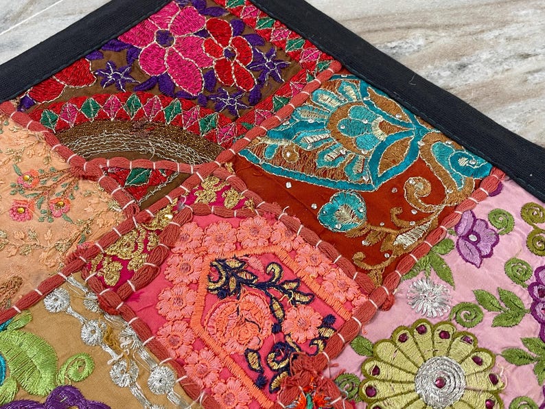 May include: Close-up of a colorful patchwork textile with a black border. The design features embroidered squares in shades of pink, orange, teal, and brown, with floral and geometric patterns. The textile is likely a decorative item, such as a pillow cover or wall hanging.