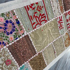 May include: A patchwork textile with a cream-colored border. The design features embroidered squares in various colors, including red, pink, blue, gold, and brown. The squares have intricate patterns and are stitched together with red thread.