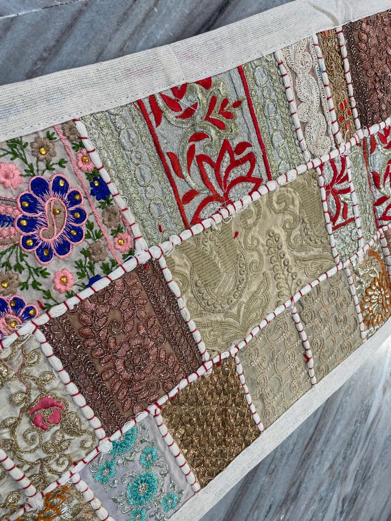 May include: A patchwork textile with a cream-colored border. The design features embroidered squares in various colors, including red, pink, blue, gold, and brown. The squares have intricate patterns and are stitched together with red thread.