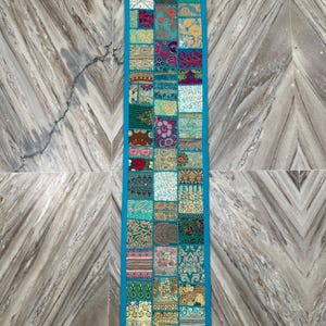 May include: A rectangular patchwork table runner with a turquoise border and tassels. The runner is composed of various fabric squares in shades of teal, pink, gold, and green. The design features floral and geometric patterns.