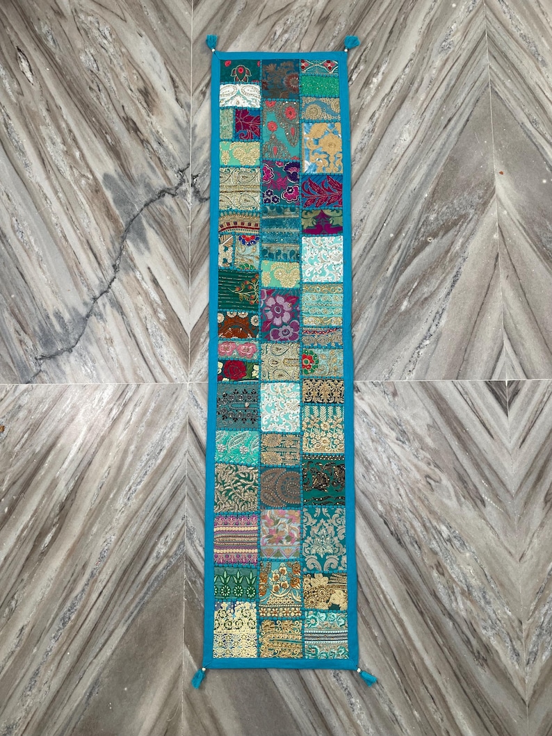 May include: A rectangular patchwork table runner with a turquoise border and tassels. The runner is composed of various fabric squares in shades of teal, pink, gold, and green. The design features floral and geometric patterns.