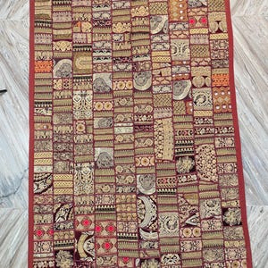 May include: A rectangular patchwork textile with a red border. The design features numerous rectangular and square patches in shades of burgundy, gold, and cream, each with intricate patterns. The overall aesthetic is ornate and detailed, suitable for home decor.