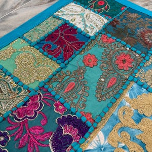 May include: A patchwork textile with a vibrant mix of colors and patterns. The design features squares of teal, turquoise, gold, pink, and burgundy fabrics, each with intricate embroidered details. A bright blue border frames the piece.