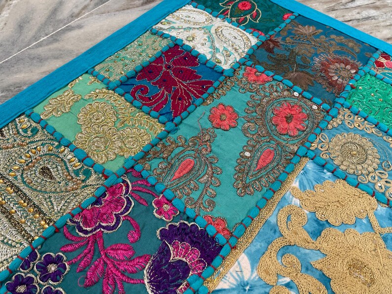 May include: A patchwork textile with a vibrant mix of colors and patterns. The design features squares of teal, turquoise, gold, pink, and burgundy fabrics, each with intricate embroidered details. A bright blue border frames the piece.