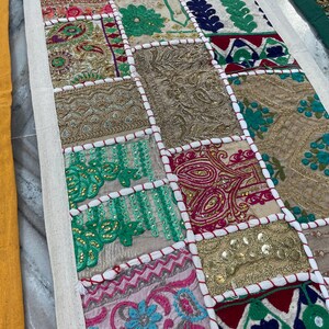 May include: A colorful patchwork textile with a variety of embroidered squares. The squares feature floral and geometric patterns in shades of gold, pink, green, and blue. The textile is bordered with a cream-colored fabric.