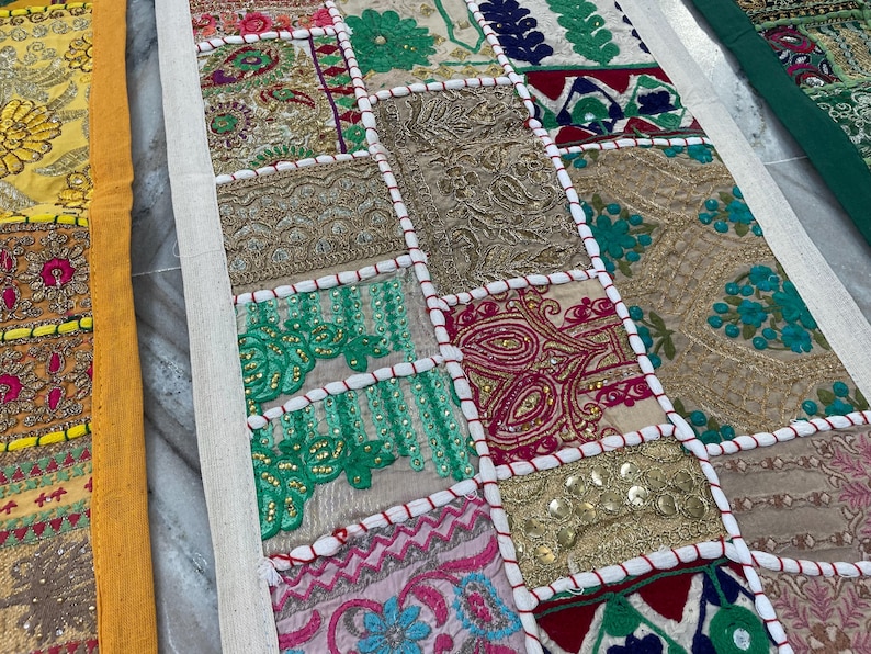 May include: A colorful patchwork textile with a variety of embroidered squares. The squares feature floral and geometric patterns in shades of gold, pink, green, and blue. The textile is bordered with a cream-colored fabric.