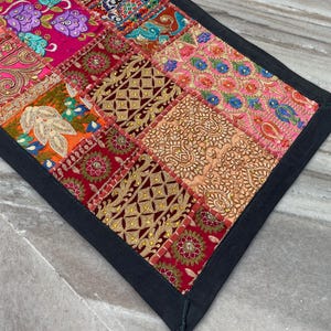 May include: A colorful patchwork textile with a black border. The design features a variety of patterned squares in shades of pink, red, orange, and blue. The textile has an intricate, handcrafted appearance.