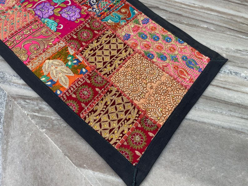 May include: A colorful patchwork textile with a black border. The design features a variety of patterned squares in shades of pink, red, orange, and blue. The textile has an intricate, handcrafted appearance.