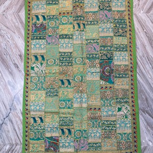 May include: A rectangular patchwork textile with a green border. The textile is composed of numerous squares in shades of green, gold, and teal, featuring various patterns and designs. The border is a solid green with a decorative trim.