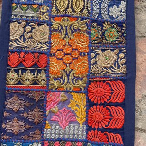 May include: A patchwork textile with a dark blue border. The design features embroidered squares in various colors, including gold, orange, red, and blue. Floral and geometric patterns are prominent in the detailed stitching.