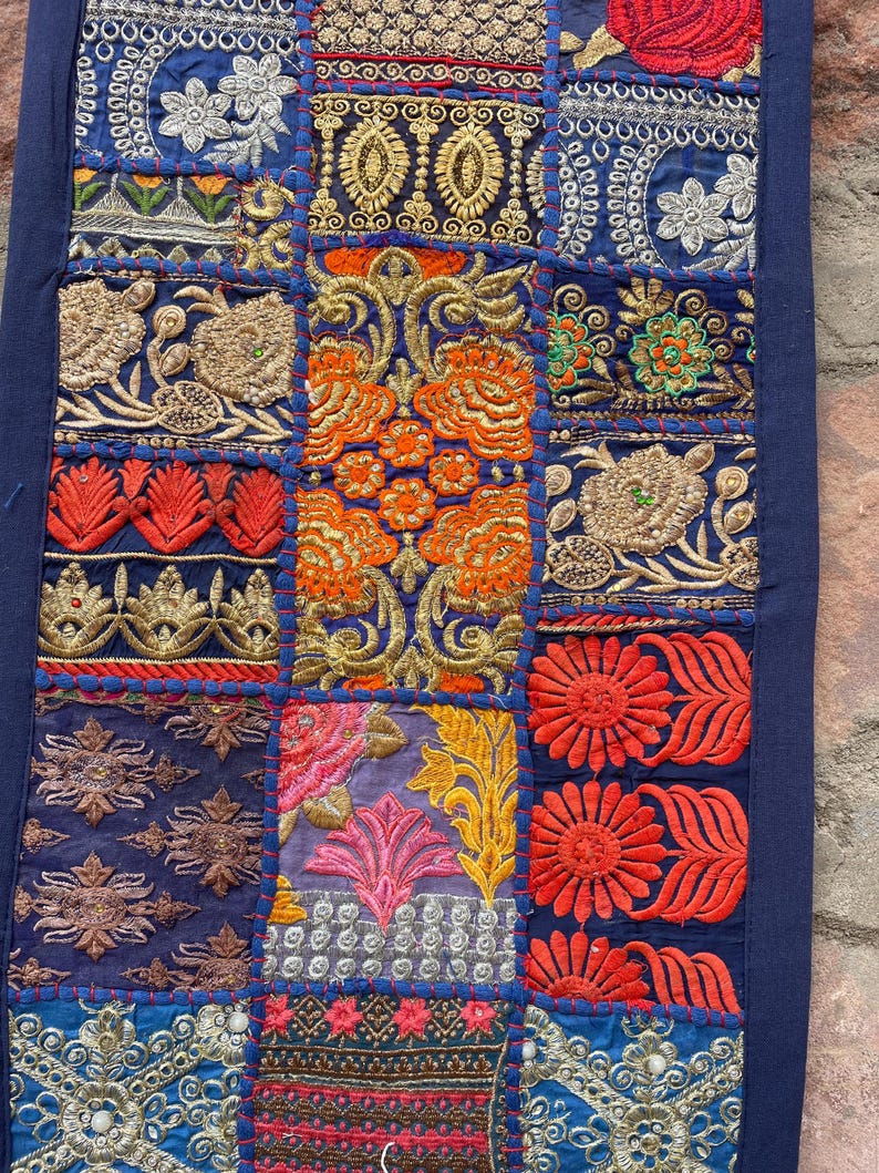 May include: A patchwork textile with a dark blue border. The design features embroidered squares in various colors, including gold, orange, red, and blue. Floral and geometric patterns are prominent in the detailed stitching.
