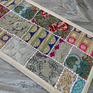 May include: A rectangular patchwork table runner with a beige border. The runner features a variety of embroidered fabric squares in shades of gold, blue, red, pink, and green. The design includes floral and geometric patterns.