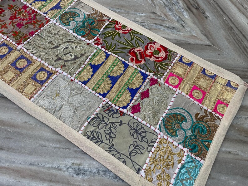 May include: A rectangular patchwork table runner with a beige border. The runner features a variety of embroidered fabric squares in shades of gold, blue, red, pink, and green. The design includes floral and geometric patterns.