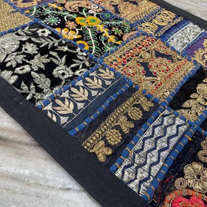 May include: A patchwork textile with a black border. The design features squares of various fabrics, including black velvet with silver embroidery, gold brocade, and floral patterns. The colors include gold, blue, black, and red.