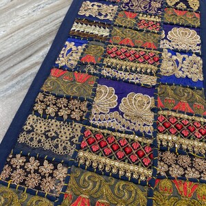 May include: A rectangular, patchwork textile with a dark blue border. The patchwork features squares of various fabrics in shades of blue, gold, red, and green, adorned with intricate embroidery and embellishments. The textile is likely a decorative item.