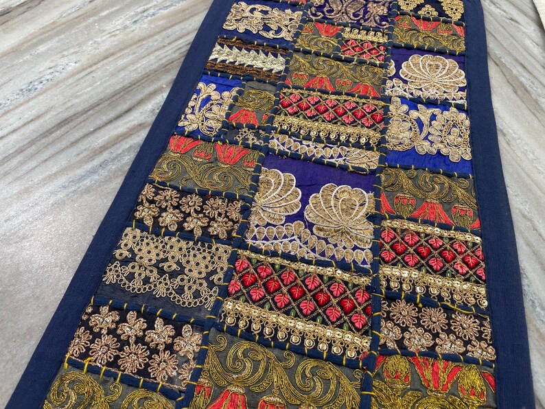 May include: A rectangular, patchwork textile with a dark blue border. The patchwork features squares of various fabrics in shades of blue, gold, red, and green, adorned with intricate embroidery and embellishments. The textile is likely a decorative item.