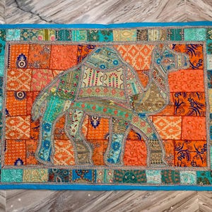 May include: A rectangular, patchwork textile featuring a camel design. The camel is composed of various patterned fabrics in shades of blue, green, and orange. The background is a patchwork of orange squares, framed by a turquoise border.