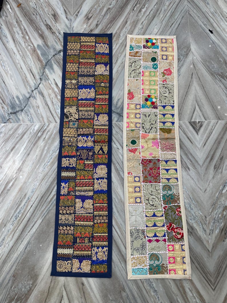 May include: Two rectangular, decorative textile runners. One is navy blue with a patchwork design of gold, red, and green embroidered squares. The other is cream-colored with a similar patchwork design featuring various colors and patterns.