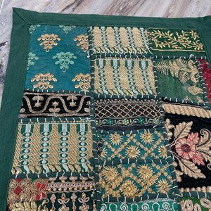 May include: A patchwork textile with a dark green border. The design features various squares with embroidered patterns in teal, gold, and dark green. Some squares have floral and geometric designs. The textile is likely used as a decorative item.