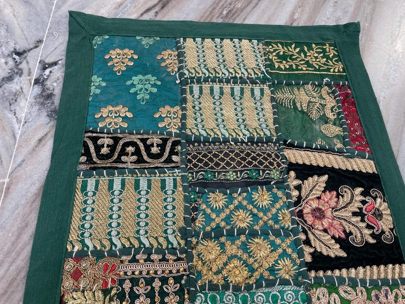 May include: A patchwork textile with a dark green border. The design features various squares with embroidered patterns in teal, gold, and dark green. Some squares have floral and geometric designs. The textile is likely used as a decorative item.
