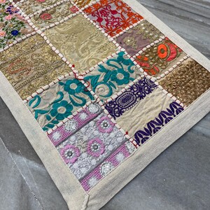 May include: A patchwork table runner with a variety of embroidered squares in gold, pink, orange, teal, and purple. The squares are bordered with white fabric and red stitching. The runner has a neutral-colored border.