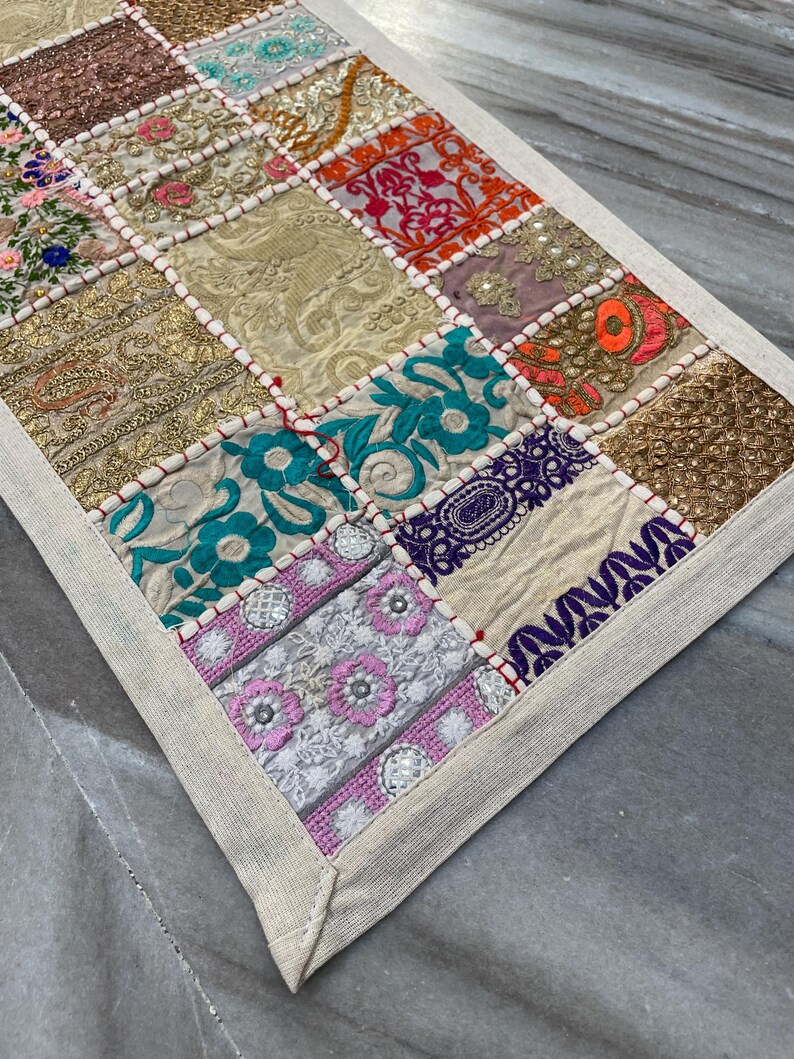 May include: A patchwork table runner with a variety of embroidered squares in gold, pink, orange, teal, and purple. The squares are bordered with white fabric and red stitching. The runner has a neutral-colored border.