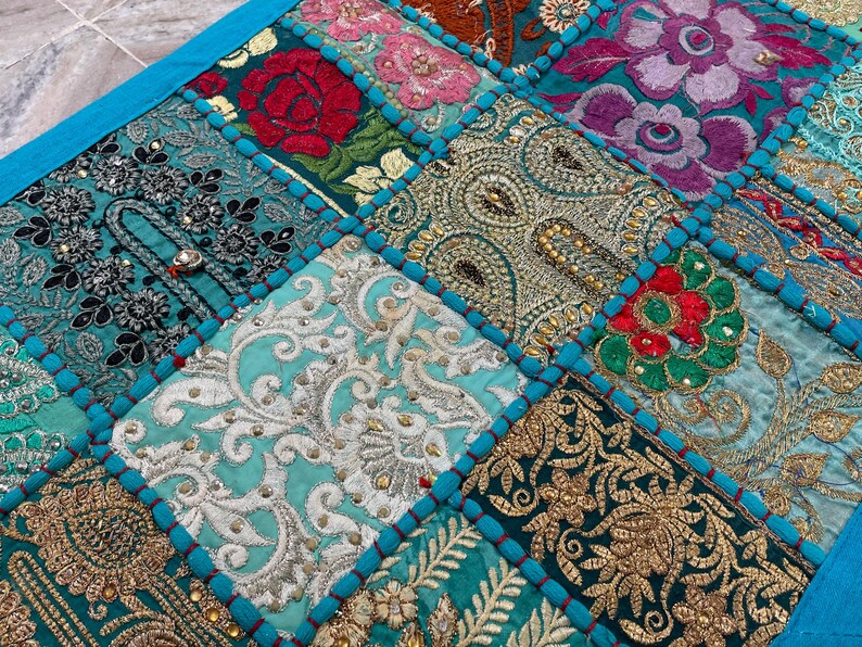 May include: A patchwork textile with a vibrant mix of colors and patterns. The design features squares of embroidered fabric in teal, turquoise, gold, and magenta. Each square has a unique floral or paisley design, with a teal border.