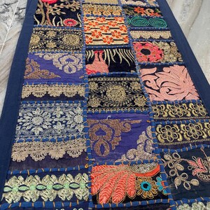 May include: A rectangular patchwork textile with a dark blue border. The patchwork features various embroidered squares in shades of blue, gold, orange, and pink. The textile is likely a table runner or decorative piece.