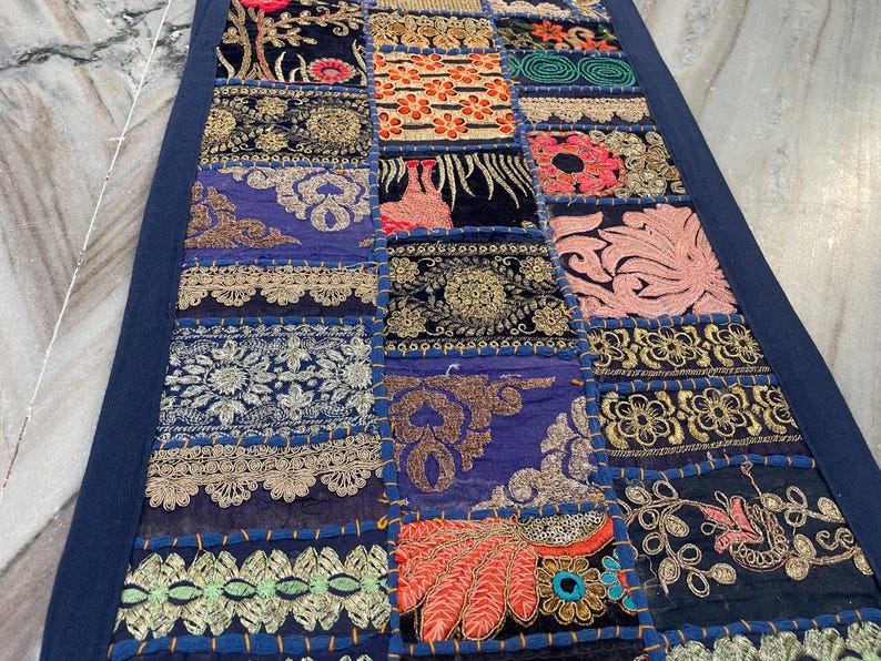 May include: A rectangular patchwork textile with a dark blue border. The patchwork features various embroidered squares in shades of blue, gold, orange, and pink. The textile is likely a table runner or decorative piece.